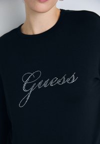 Nahaufnahme einer Person, die ein schwarzes Shirt trägt, auf dem "Guess" in silbernen Strasssteinen über der Brust steht.