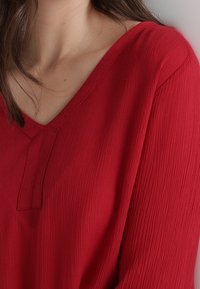 Blusa rossa, strutturata, con scollatura a V e maniche lunghe. Presenta un delicato motivo increspato e un piccolo inserti centrale.