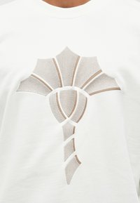 Felpa bianca con un grande motivo ritagliato a forma di conchiglia, con uno sfondo in tessuto beige testurizzato e contornata da una sottile linea marrone.