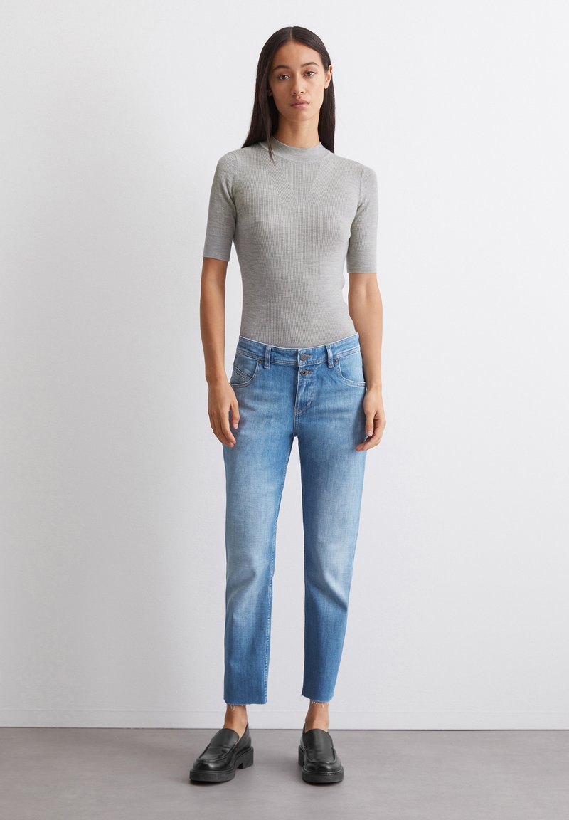 Graues, geripptes T-Shirt mit kurzen Ärmeln, kombiniert mit hellblauen, gerade geschnittenen Jeans mit Knopfverschluss, Fünf-Taschen-Design und fransigem Saum. Schwarze Loafer.