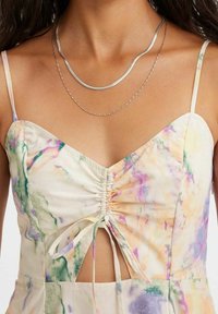 Robe multicolore avec un nœud devant et un corsage froncé, présentant des nuances de vert, violet et jaune. Bretelles fines, texture de tissu lisse.