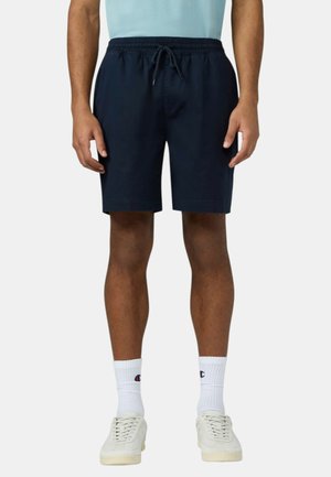 Uomo che indossa pantaloncini blu navy con coulisse, calzini bianchi con il logo Champion e scarpe da ginnastica bianche, in piedi con le braccia rilassate ai lati.