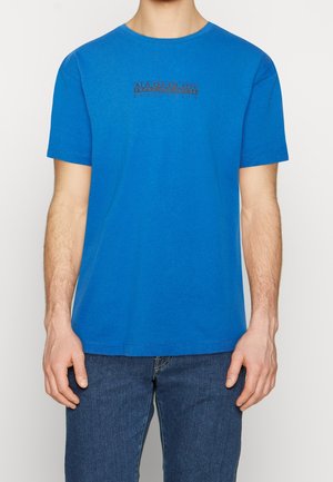 Uomo che indossa una maglietta blu a maniche corte con il logo "Napapijri geographic" e jeans blu, di fronte con le mani rilassate ai lati.