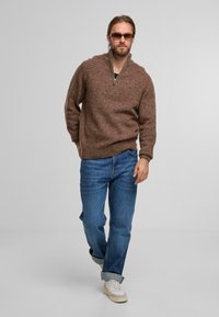 Braun gemusterter Strickpullover mit einem halben Reißverschlusskragen, kombiniert mit blauen, locker sitzenden Jeans und weißen Sneakers. Das Model steht vor einem schlichten Hintergrund.
