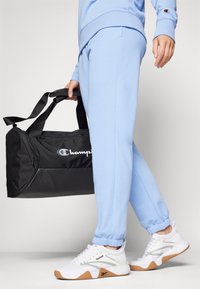 Sac de sport noir avec logo embossé, associé à un pantalon de survêtement bleu clair avec des poignets et des chaussures blanches avec des semelles en caoutchouc et des accents argentés.