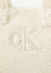 Tela de borrego color crema que presenta un logotipo bordado "CK" en un tono a juego. La textura parece suave y esponjosa.