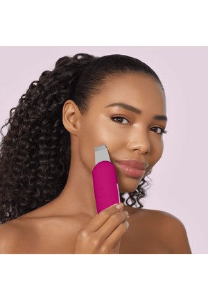 Woman with curly hair using a pink facial cleansing device on her cheek against a light purple background.