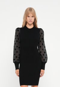 Abito nero con texture a coste e lunghe maniche trasparenti che presentano pois neri. Collo solido e silhouette aderente.