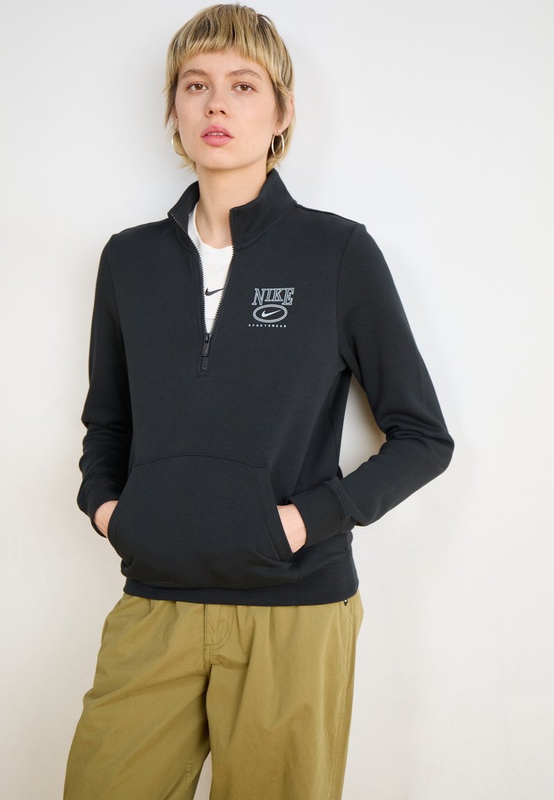 Sudadera negra con cremallera hecha de una tela suave, con un bolsillo frontal y un logo de Nike en el pecho. Combinada con pantalones verde oliva.