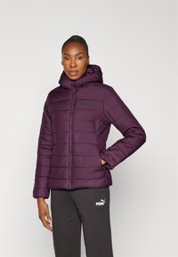 Giacca a vento viola con cappuccio, caratterizzata da sezioni trapuntate e un logo Puma nero. Indossata con pantaloni grigi, che mostrano un fit comodo.