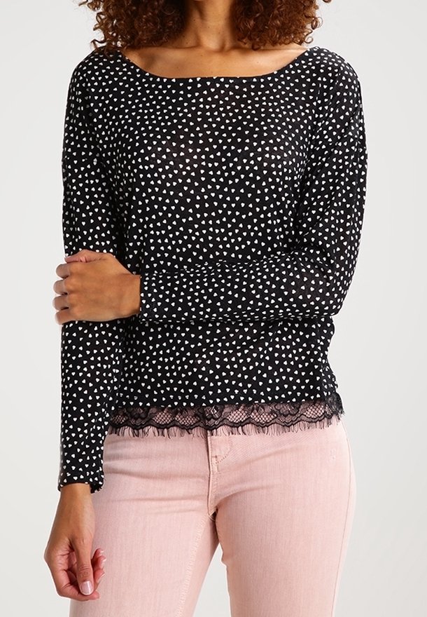 Kobieta w czarnej bluzce z długim rękawem, ozdobionej małymi białymi wzorami serc oraz koronkowym wykończeniem, zestawiona z jasnoróżowymi spodniami.