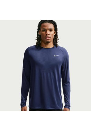 Man met dreadlocks die een lange mouw navyblauw Nike sportshirt draagt en recht vooruit kijkt tegen een effen achtergrond.
