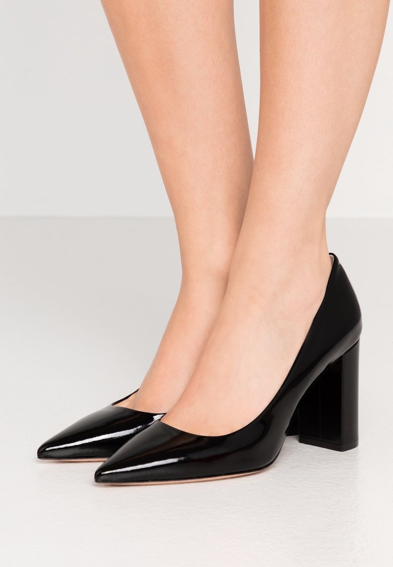 Jambes portant des chaussures en cuir verni noir brillant à bout pointu et talon bloc sur un fond clair.