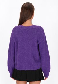 Pulover fuzos mov cu mâneci lungi, tip balon, și tiv din material cu nervuri. Materialul texturat adaugă căldură, fiind asortat cu o fustă plisată neagră.