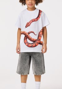 Weißes Baumwoll-T-Shirt mit einem großen rot-musternden Schlangendesign, kombiniert mit grauen Jeansshorts und weißen Knöchel-Socken.