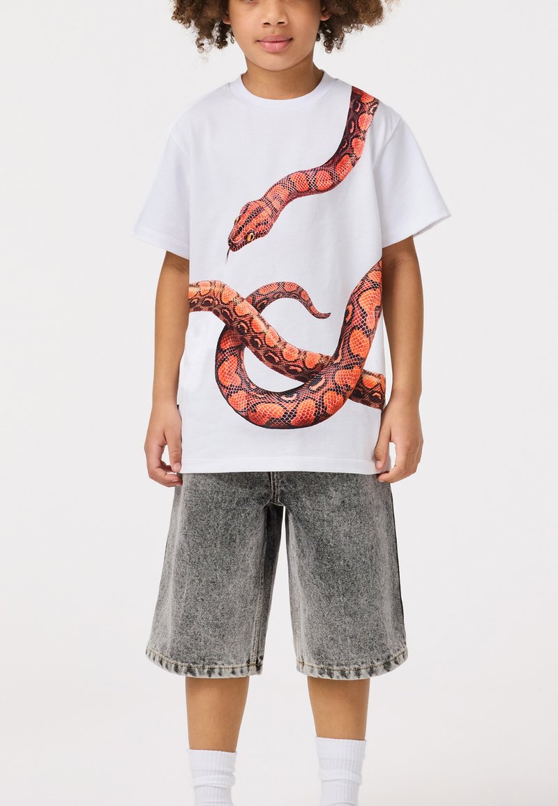 Weißes Baumwoll-T-Shirt mit einem großen rot-musternden Schlangendesign, kombiniert mit grauen Jeansshorts und weißen Knöchel-Socken.