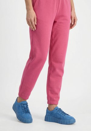 Osoba stojící před bílým pozadím, oblečená v jasně růžových teplácích a modrých platformových teniskách.