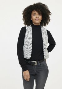 Gestrickte Weste im weißen und grauen Fischgrätenmuster, über einem schwarzen Rollkragenpullover getragen. Verfügt über ein tailliertes Design und einen offenen Vorderteil. Schwarzer Gürtel.