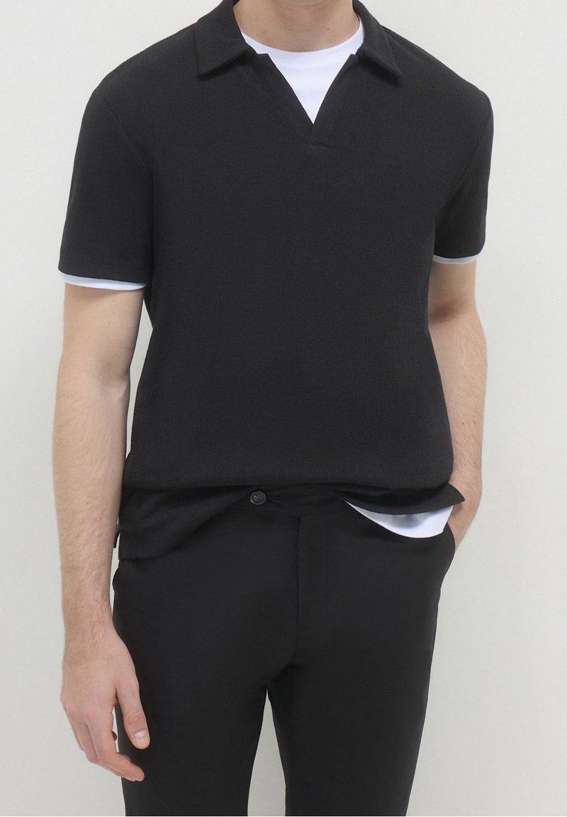 Homme portant une chemise noire à manches courtes avec col superposée à un T-shirt blanc, associée à un pantalon noir, main dans la poche, debout devant un fond uni.