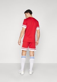 Röd idrottströja och shorts med vita ränder; modellen har på sig vita strumpor med blå band och fotbollsskor, sedd bakifrån.