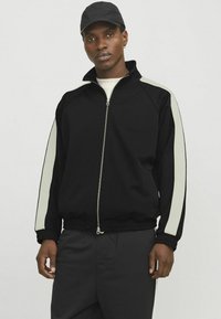 Sort zip-up træningsjakke med lysegrå side-striber, ribbede manchetter og elastisk talje. Bæres med en lysfarvet skjorte og kasket.