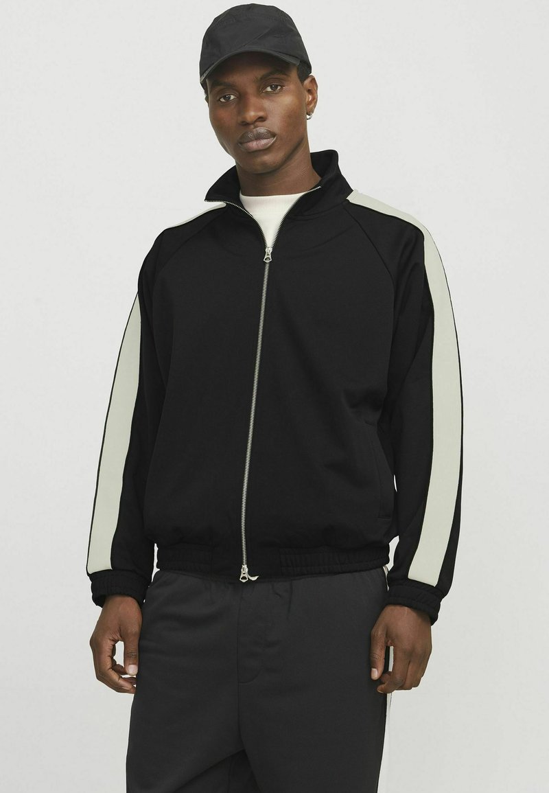 Sort zip-up træningsjakke med lysegrå side-striber, ribbede manchetter og elastisk talje. Bæres med en lysfarvet skjorte og kasket.