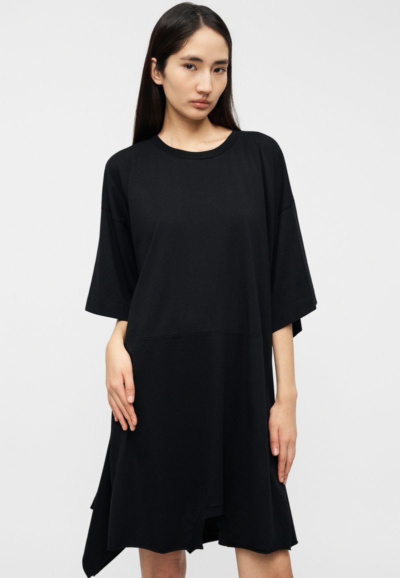 Robe t-shirt noire oversize en coton doux, avec manches courtes, col rond, et ourlet asymétrique avec fentes latérales.