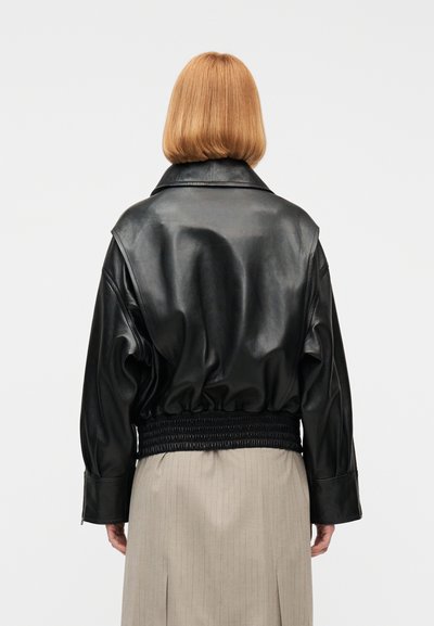 Άτομο με ίσια κοκκινωπά μαλλιά μέχρι τους ώμους, φοράει μαύρο δερμάτινο μπουφάν με ζωνάκι στη μέση και μπεζ φούστα με λεπτές ρίγες, γυρισμένο προς τα πίσω.