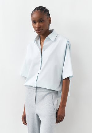 Kobieta z fryzurą cornrow, ubrana w luźną jasnoniebieską koszulkę z krótkim rękawem, wpuszczoną w teksturowane jasnoszare spodnie, stoi na białym tle.