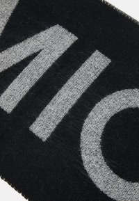 Černá textilie s velkým šedým textem. Tkanina má měkkou, strukturální vazbu, s roztrhanými okraji a jemným leskem.