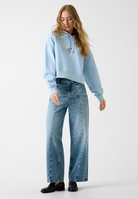 Hoodie crop bleu clair avec capuche à cordon et logo, associé à un jean en denim déchiré à taille haute et jambes larges, ainsi que des mocassins noirs.