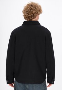 Giacca a camicia nera con colletto, maniche lunghe e realizzata in un tessuto testurizzato. Presenta polsini con bottone e orlo arrotondato, abbinata a pantaloni in denim blu.