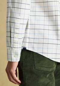 Weiß kariertes Hemd mit blauen und schwarzen Streifen, Knopfmanschetten. Kombiniert mit dunkelgrünen Cordhosen mit einer Gesäßtasche.