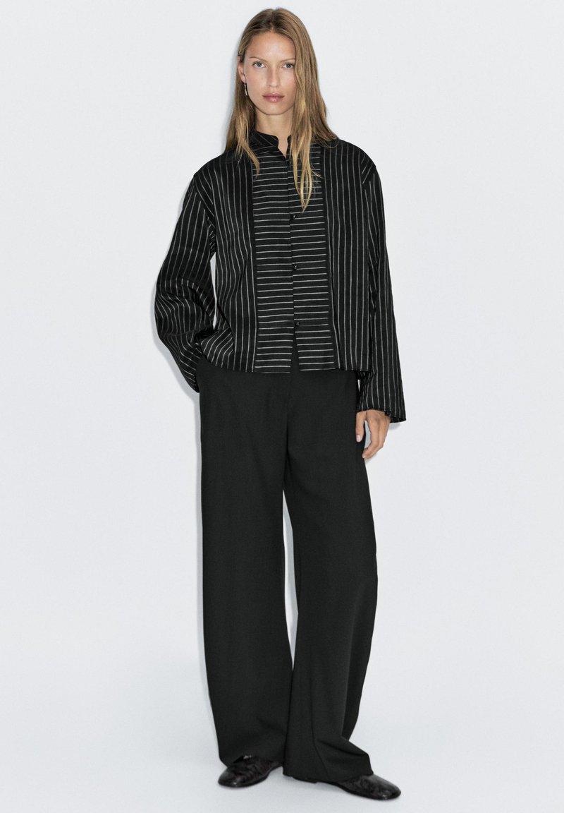 Zwarte gestreepte button-up jas met een mandarin kraag, gecombineerd met wijdvallende zwarte broeken. Gladde stof met verticale en horizontale lijnen.