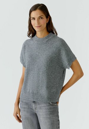 Eine Frau mit braunen Haaren, die einen kurzärmligen grauen Strickpullover und hellgraue Jeans trägt, steht mit einer Hand in der Tasche vor einem schlichten Hintergrund.