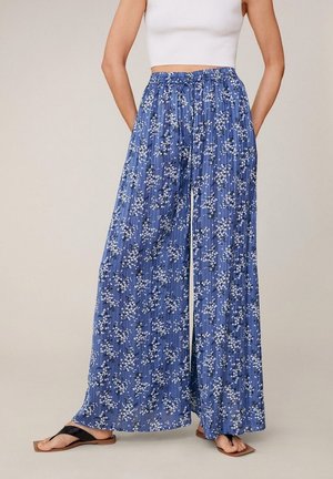 Femme portant un pantalon large bleu à motif floral blanc, associé à un crop top blanc et des sandales noires, debout les mains dans les poches.