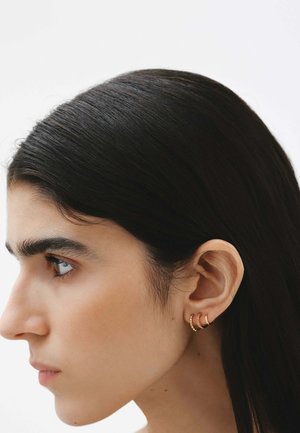 Pendientes de aro dorados exhibidos en la oreja derecha, con un diseño retorcido que presenta tres tamaños distintos apilados juntos. Acabado suave, estilo minimalista.