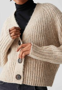 Inquadratura ravvicinata di una persona che abbottona un cardigan lavorato a maglia beige su un dolcevita nero, indossando jeans scuri.