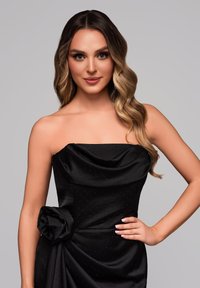 Černé saténové šaty bez ramínek s drapovaným korzátem a květinovými ozdobami na boku. Tkanina obsahuje jemné tečky, které odrážejí světlo.