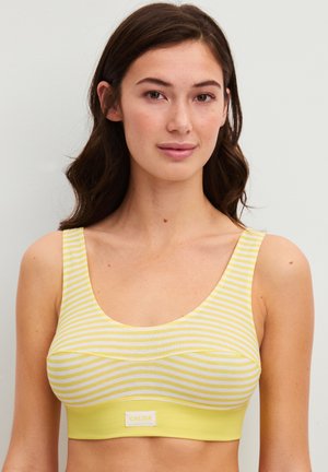 Jonge vrouw met donkerbruin haar draagt een geel-wit gestreept mouwloos topje tegen een effen neutrale achtergrond.