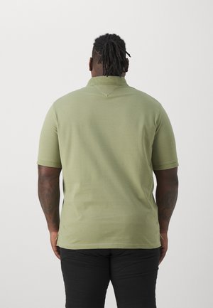 Man met korte dreadlocks met de rug naar de camera, draagt een lichtgroen poloshirt en zwarte broek, tegen een effen witte achtergrond.