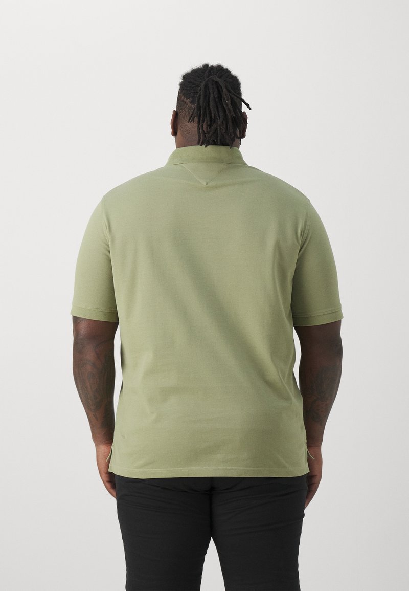 Homme avec de courtes dreadlocks de dos, portant un polo vert clair et un pantalon noir, debout devant un fond blanc uni.