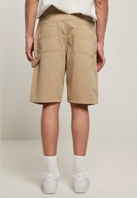 Beige cargo šortky s mnoha kapsami, vyrobené z lehkého materiálu. Mají zesílené švy a volný střih, nošeny s bílými teniskami.