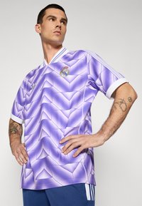 Camiseta de fútbol con un patrón morado y blanco que presenta un diseño en zigzag, un cuello en V y los logotipos de Adidas y Real Madrid.