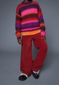 Sweter z dzianiny w wielokolorowe poziome paski (różowy, fioletowy, czerwony, pomarańczowy), w połączeniu z szerokimi czerwonymi spodniami z korduroju i masywnymi czarnymi butami z białymi futrzanymi akcentami.