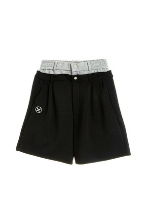 Joven con rastas lleva puesta una camiseta blanca y roja de gran tamaño con motivos de Japón, pantalones cortos negros, calcetines negros y botas de trabajo marrones sobre fondo blanco.