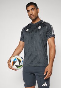 Jersey deportivo gris oscuro con un patrón texturizado, logo y acentos blancos, combinado con pantalones cortos grises. El sujeto sostiene un balón de fútbol multicolor.