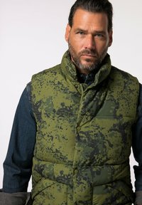 Gilet imbottito verde con motivo camouflage e colletto alto, dotato di bottoni a pressione e design trapuntato. Indossato sopra una camicia di denim.