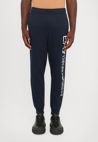 Navy sweatpants with elastic waistband, tapered legs, and white branding along the side. Paired with black sneakers featuring thick soles.