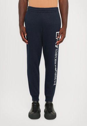 Navy sweatpants with elastic waistband, tapered legs, and white branding along the side. Paired with black sneakers featuring thick soles.